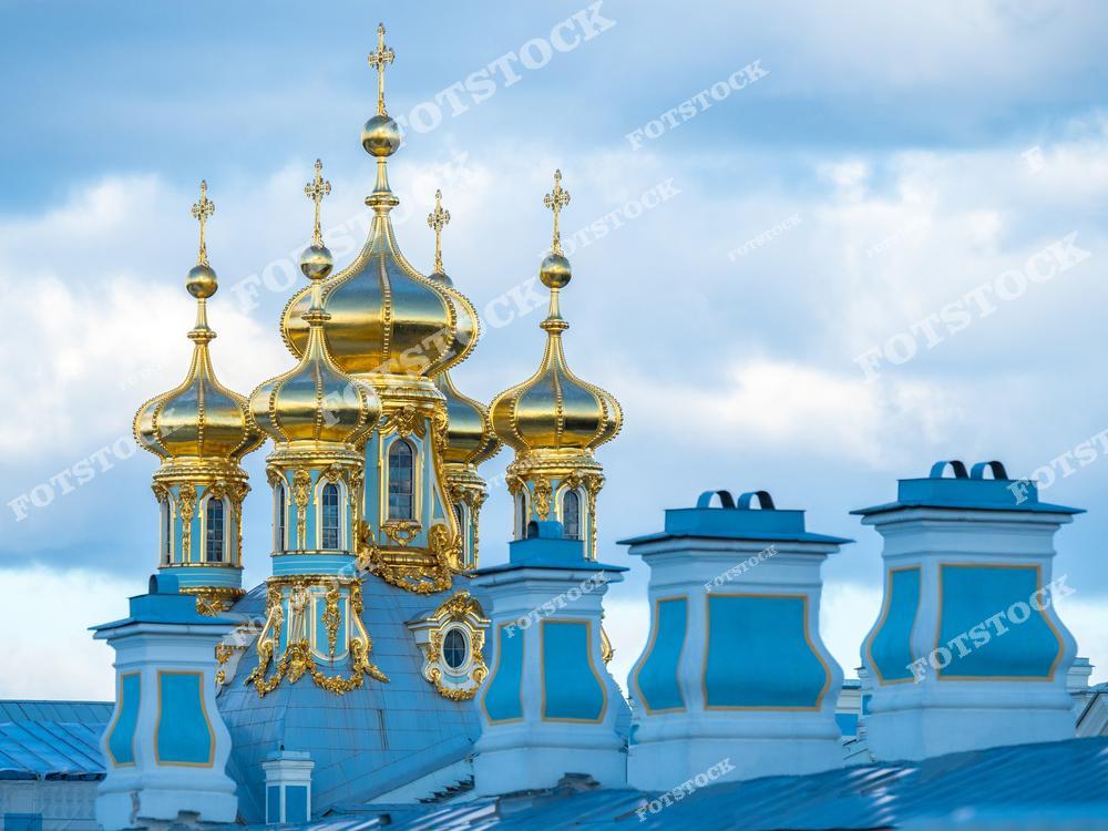 На снимке изображены купола Церкви Воскресения Христова, расположенной в Екатерининском дворце в Царском Селе. Яркий золотой декор куполов прекрасно контрастирует с голубыми оттенками неба. Летний солнечный свет подчеркивает роскошь и изящество архитектурного стиля барокко. Этот дворцово-храмовый комплекс является одной из самых значимых достопримечательностей Санкт-Петербурга и России.
